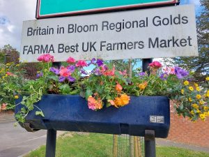 A pot of flowers under a sign reading "Britain in Bloom Regional Golds FARMA Best UK Farmers Market"